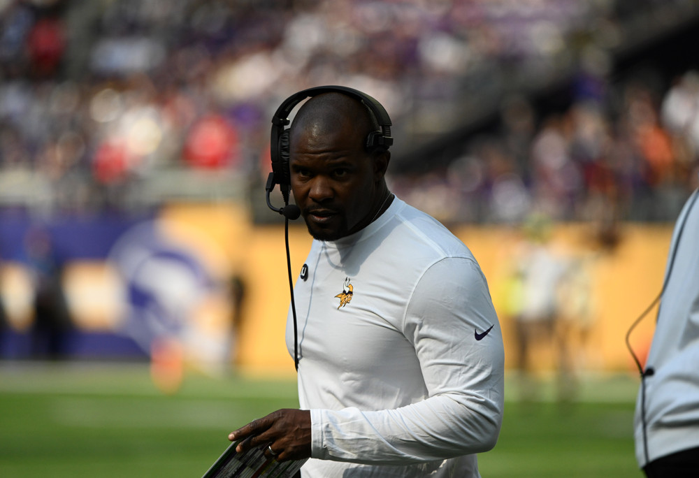 Viking coach Brian Flores, wearing a white turtleneck and a headset, stares outward along the sideline