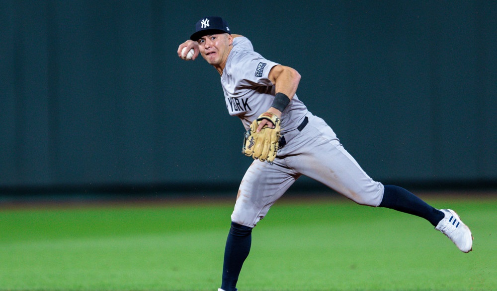 Anthony Volpe, wearing a grey uniform with blue stirrups, makes a throw to first base while raising his left leg.