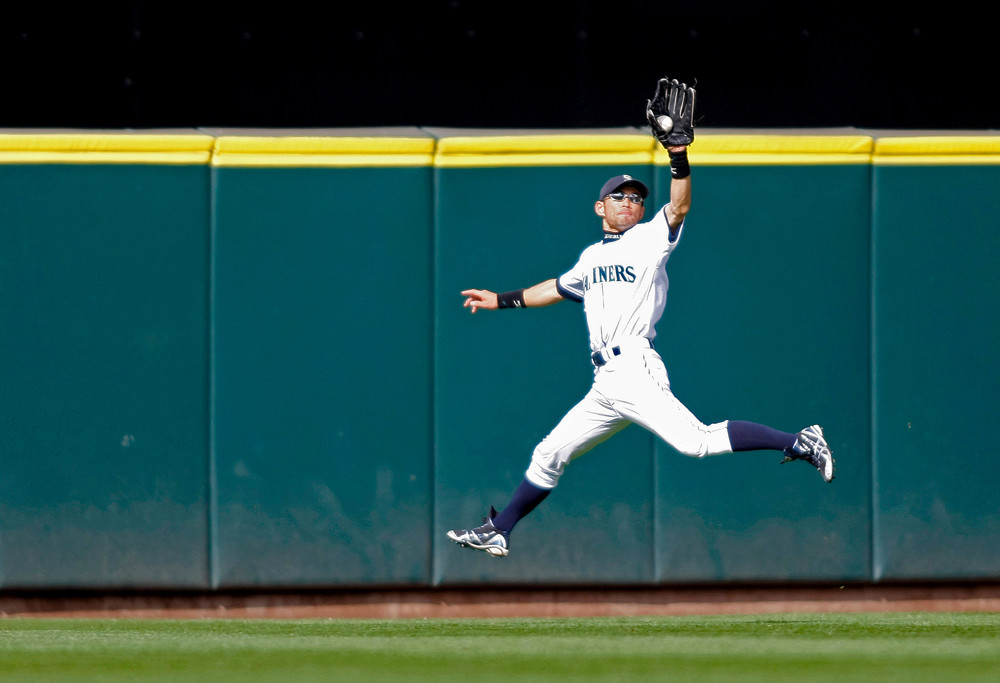 Ichiro Suzuki catches a fly ball on the run by reaching up with his glove hand as he retreats. He's wearing an all-white baseball uniform with Mariners written across the front, and blue baseball stirrups.