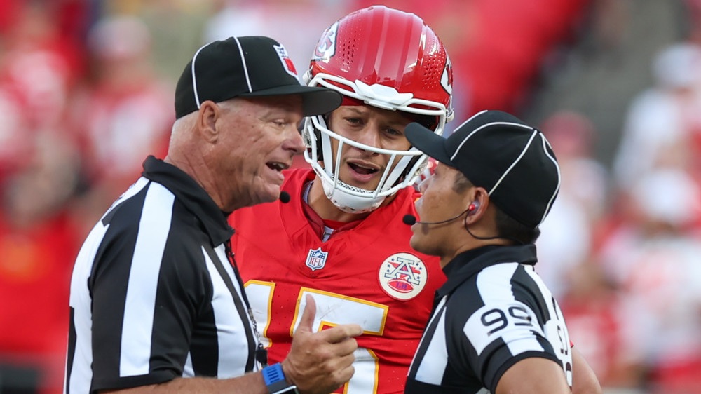 Two football officials wearing striped shirts talk while Chiefs QB Patrick Mahomes stands in front of them and listens.