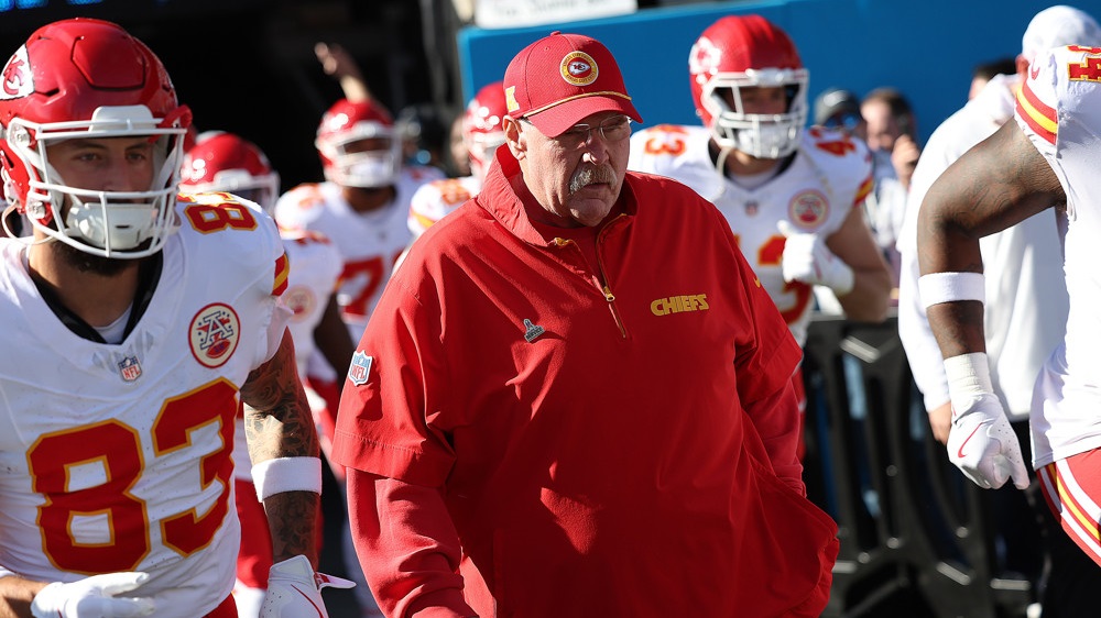 Andy Reid, wearing a red pullover jacket, comes onto the field with his team running behind him.